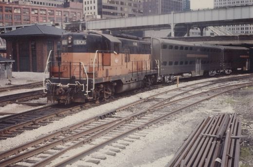 A black and orange diesel locomotive numbered 806 pulls two gray passenger cars on multiple tracks in a rail yard, with buildings and an elevated highway bridge in the background.