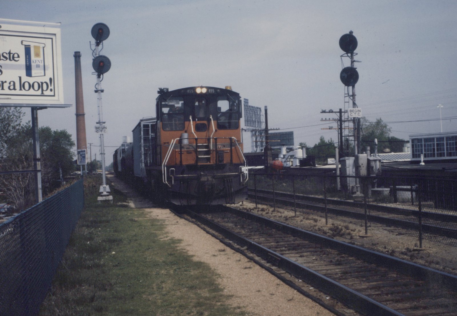 A Chicago and North Western GP9 diesel locomotive, number 455, is shown pulling freight cars through a grade crossing with signals and a billboard in the background.