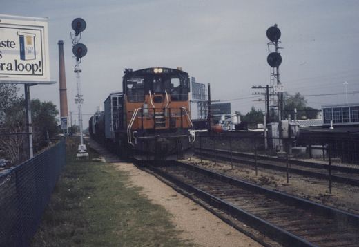 A Chicago and North Western GP9 diesel locomotive, number 455, is shown pulling freight cars through a grade crossing with signals and a billboard in the background.