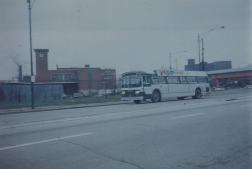 A green and white CTA bus drives down a city street under cloudy skies, with a brick building and an overpass in the background.
