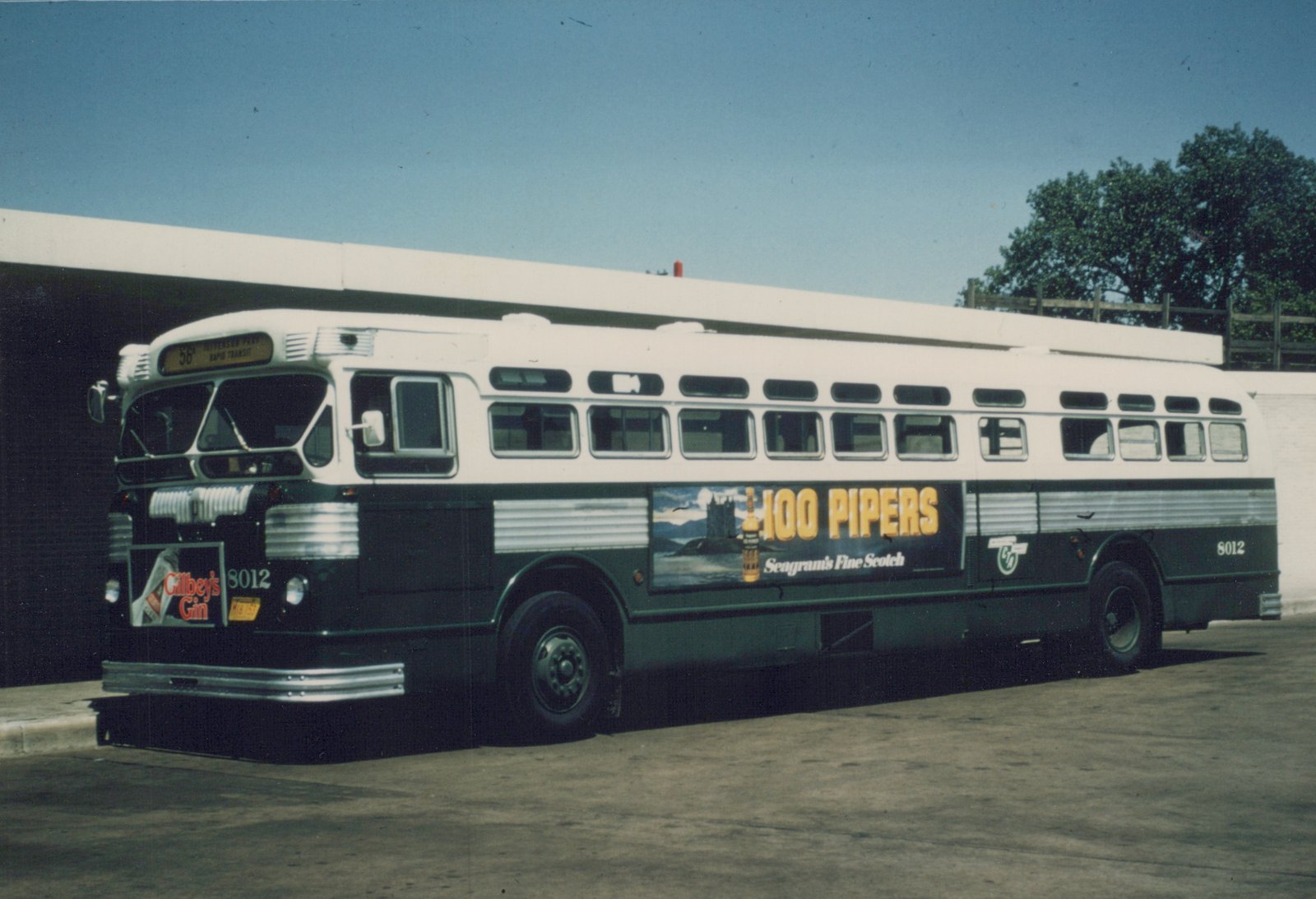 A vintage Chicago Transit Authority bus, model Flxible New Look, number 8012, is parked in a garage. The front displays route 56 to Jefferson Park and has a Gilbey's Gin ad, while the side features a large 100 Pipers whiskey advertisement. The bus is painted white and green with silver trim.