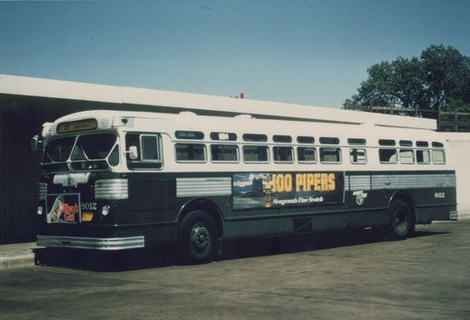 A vintage Chicago Transit Authority bus, model Flxible New Look, number 8012, is parked in a garage. The front displays route 56 to Jefferson Park and has a Gilbey's Gin ad, while the side features a large 100 Pipers whiskey advertisement. The bus is painted white and green with silver trim.