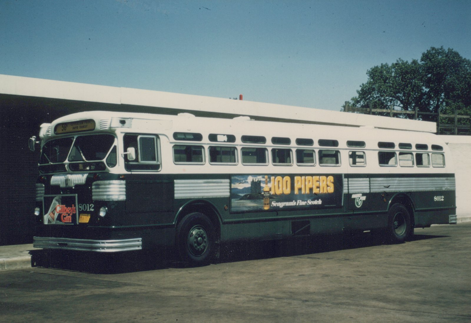 A white and green CTA bus numbered 8012 with advertisements for '100 Pipers' Scotch and 'Gilbey's Gin' is parked sideways in a paved yard under a clear blue sky.