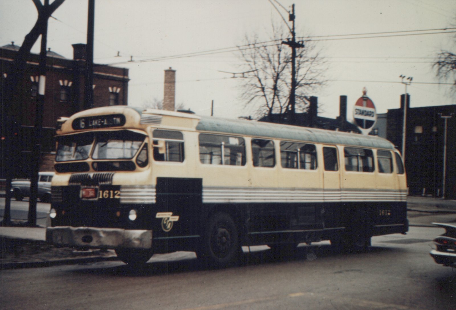 A vintage Chicago Transit Authority bus, number 1612, painted in cream and green livery, is stopped on a city street under an overcast sky with bare trees, with a Standard gas station sign in the background.