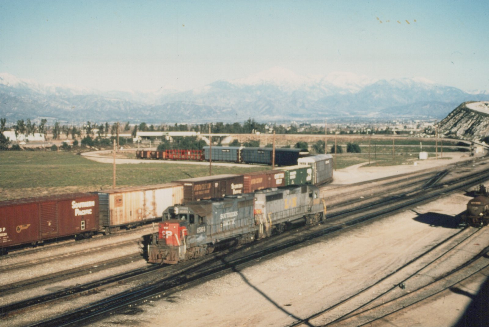 A Southern Pacific GP35 diesel locomotive, number 6564, pulls a freight train on a sunny day with snow-capped mountains in the background.