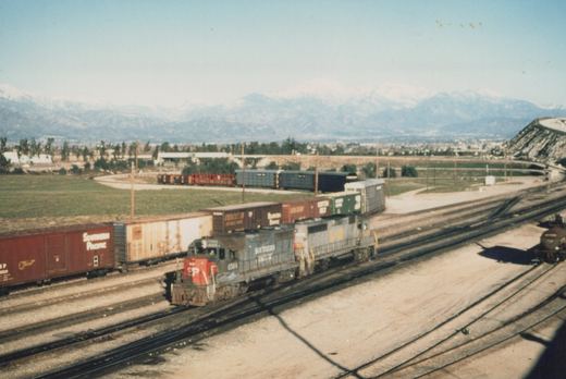 A Southern Pacific GP35 diesel locomotive, number 6564, pulls a freight train on a sunny day with snow-capped mountains in the background.
