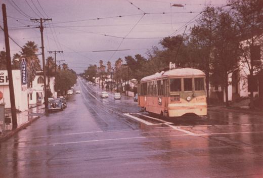A vintage green and orange streetcar with the number 1307 is stopped on a rain-slicked street, with overhead wires and a 'HUDSON' business sign visible in the background.