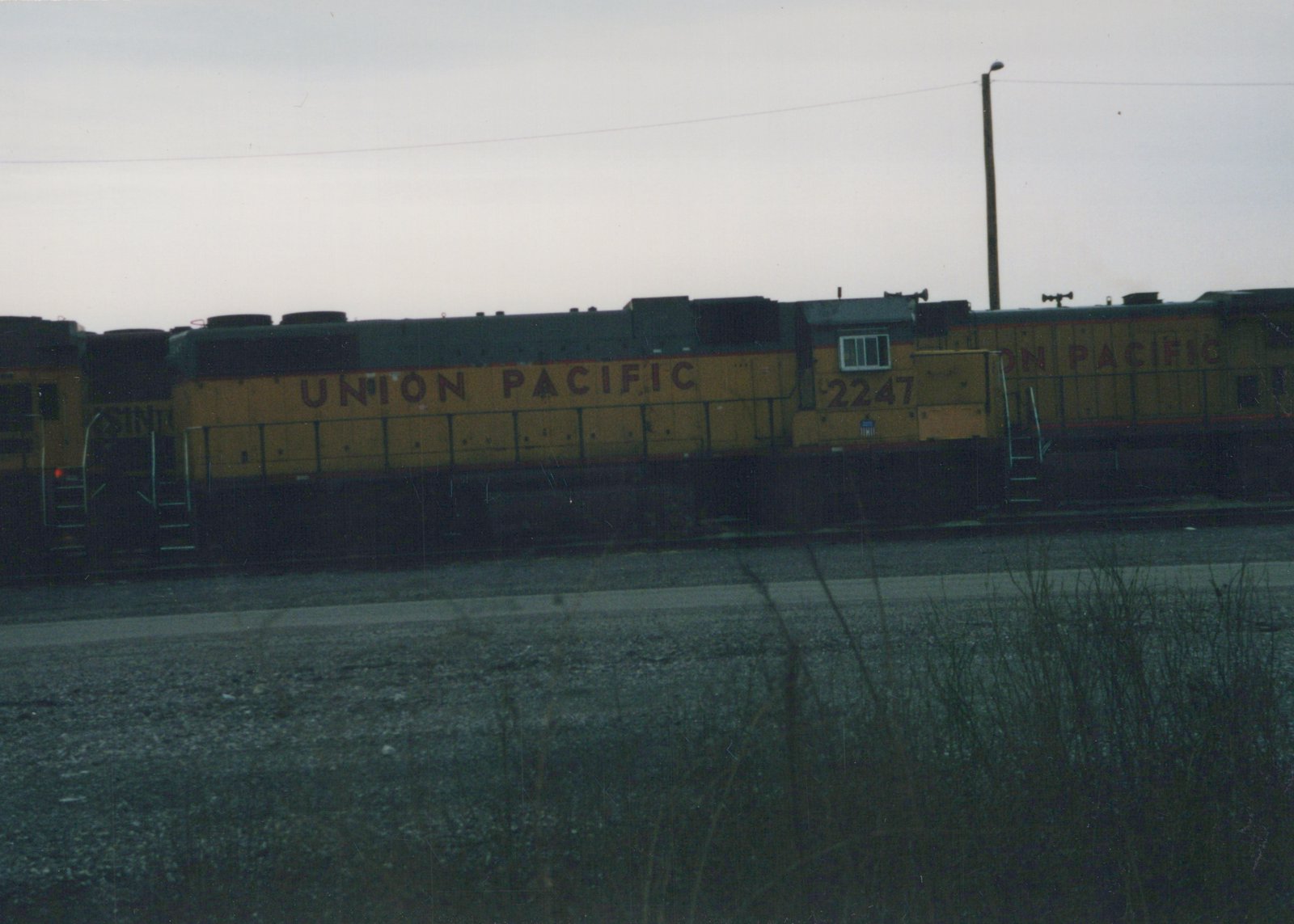 A yellow and black Union Pacific locomotive, number 2247, is parked on a railroad track in a gravel yard with dry grass on a cloudy day.
