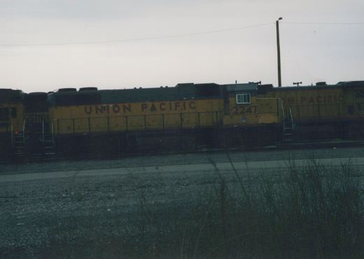 A yellow and black Union Pacific locomotive, number 2247, is parked on a railroad track in a gravel yard with dry grass on a cloudy day.