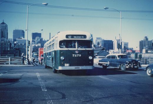 Front three-quarter view of a green and cream Chicago Transit Authority bus, number 7279, on route 42 Halsted-79, driving on a bridge with city buildings behind it.