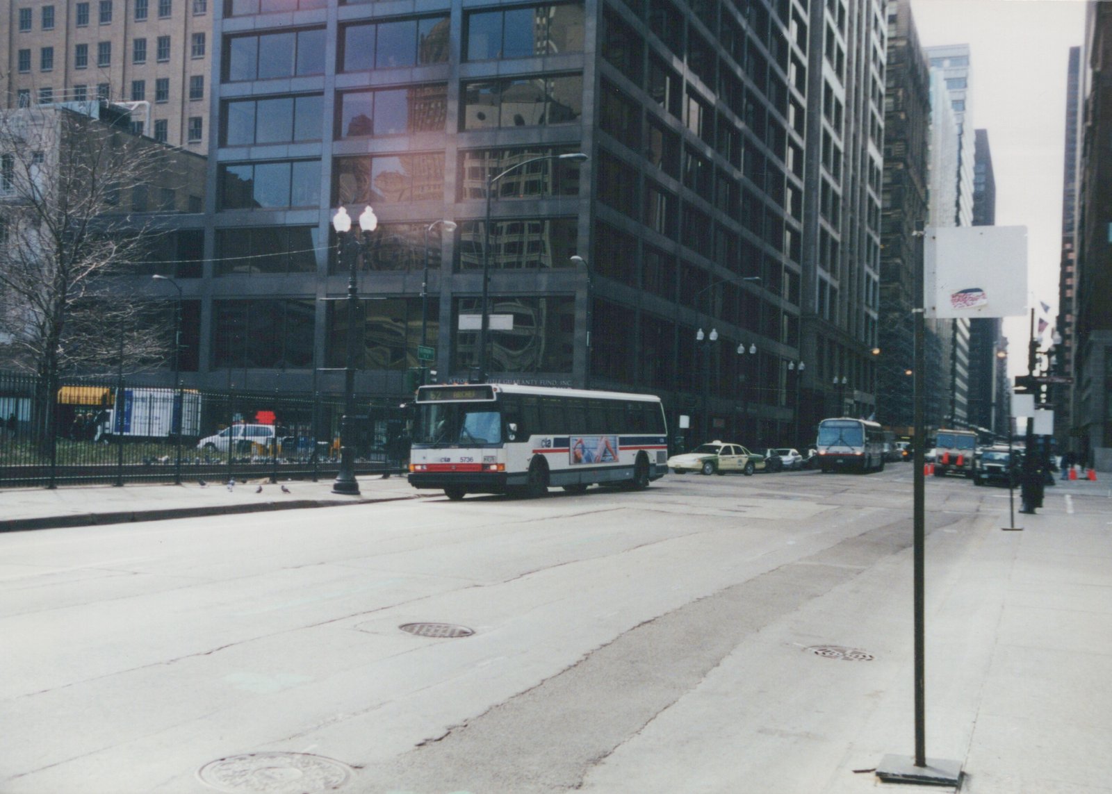 A white Chicago Transit Authority bus, number 5736, is driving down a wide city street lined with tall buildings on a cloudy day, with other vehicles and street signs visible.