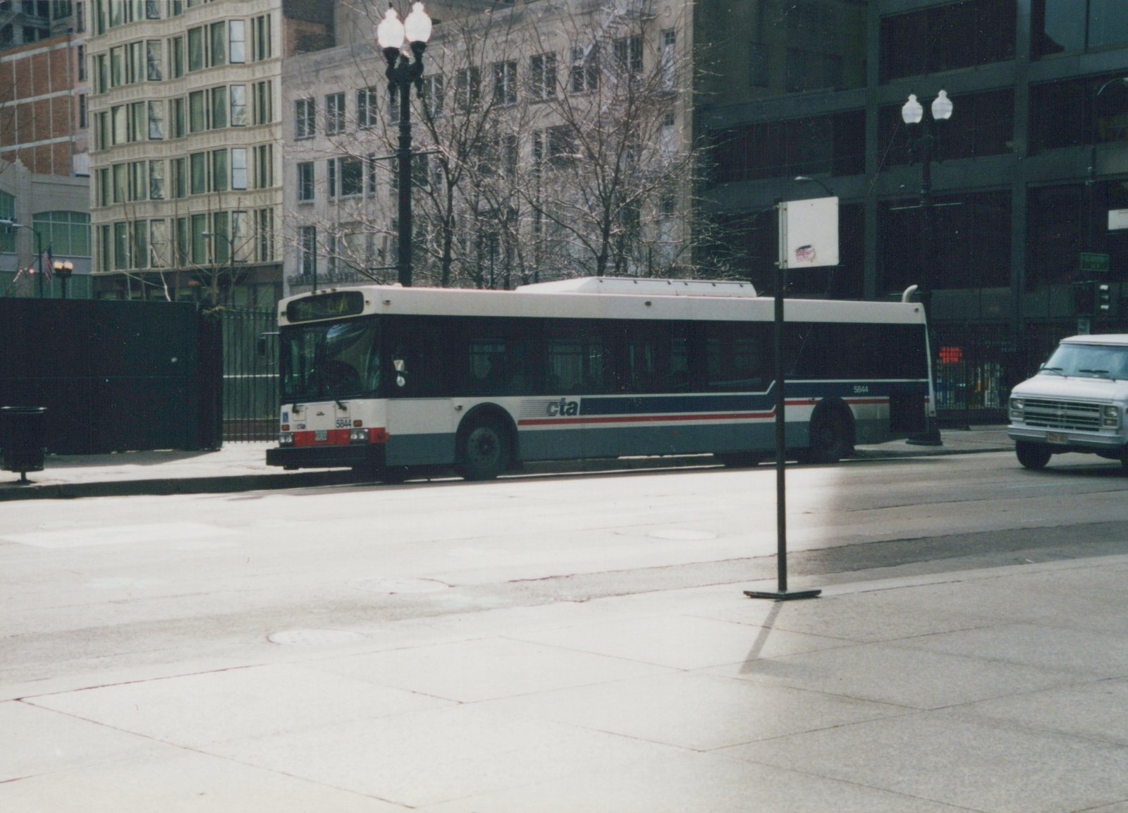 A white, blue, and red Chicago Transit Authority bus, number 5844, is parked on a city street with bare trees and snow on the ground, with a white van parked nearby.