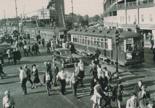 A bustling 1940s street scene in Chicago with two vintage streetcars, many pedestrians, and classic automobiles at the intersection of Clark and Addison, with Wrigley Field visible in the background.