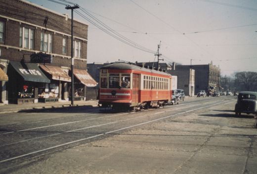 A red and cream streetcar with the number 6184 and route 81 on its destination board travels down a street lined with brick buildings and vintage automobiles.