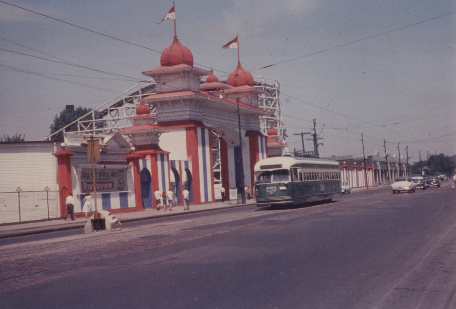 A green and cream PCC streetcar numbered 4060 travels down a street lined with power poles, passing a large red, white, and blue amusement park entrance with a roller coaster in the background.