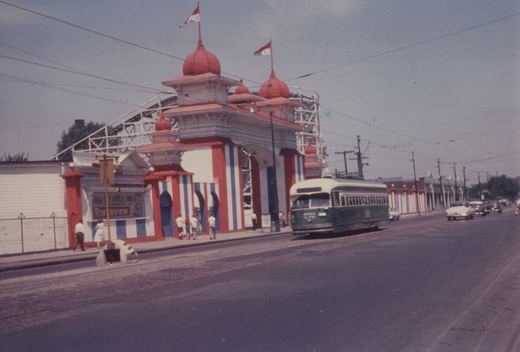 A green and cream PCC streetcar numbered 4060 travels down a street lined with power poles, passing a large red, white, and blue amusement park entrance with a roller coaster in the background.
