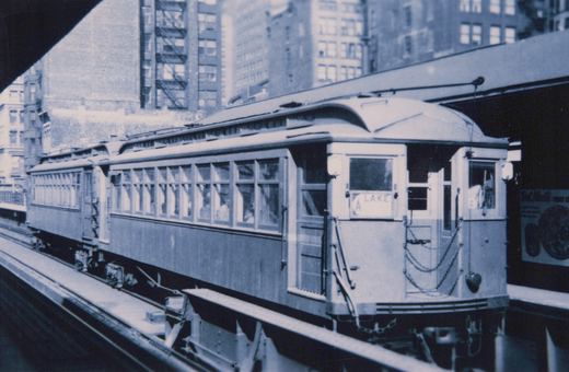 A vintage black-and-white photo of a wooden rapid transit car on an elevated track, with a destination sign reading 'A LAKE' and city buildings in the background.