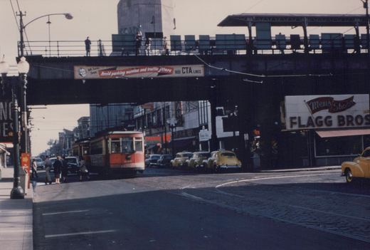 A vintage color photo showing a red and cream streetcar on a city street with cobblestones, yellow taxis parked along the curb, an elevated train platform above, and storefronts including 'Flagg Bros' and 'Hardy'.