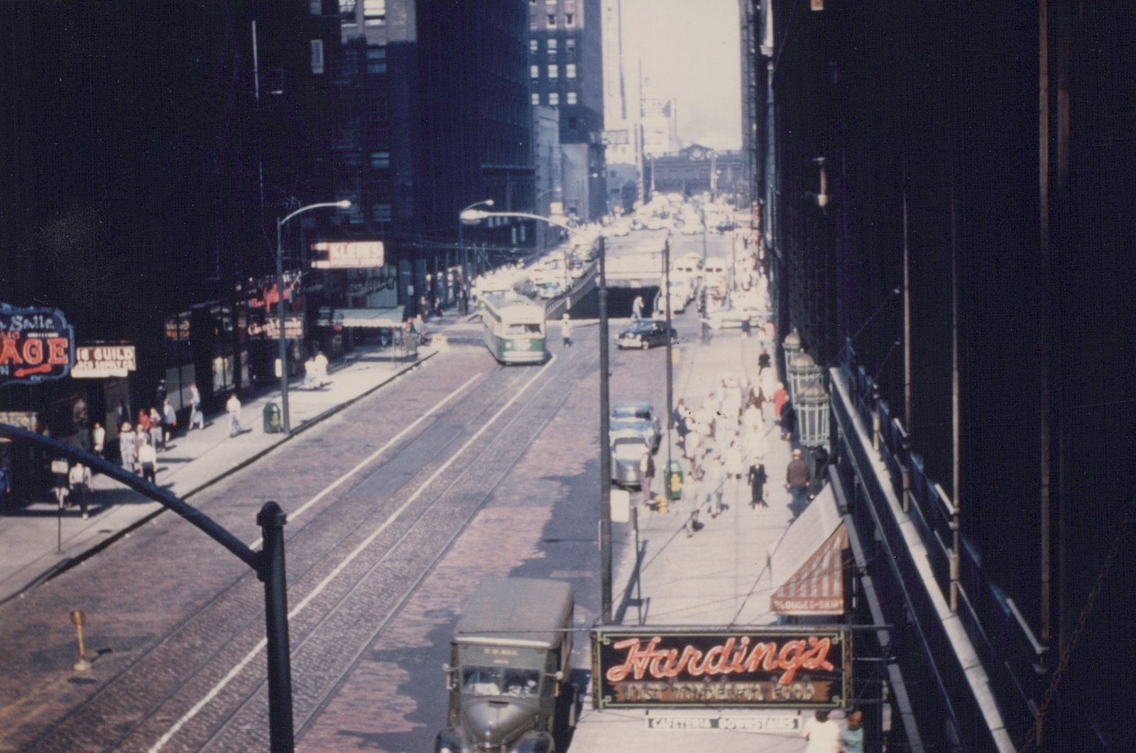 A green and cream PCC streetcar on a cobblestone street in Chicago, surrounded by pedestrians, vintage cars, and storefront signs including 'Harding's' and 'U.S. Mail'.