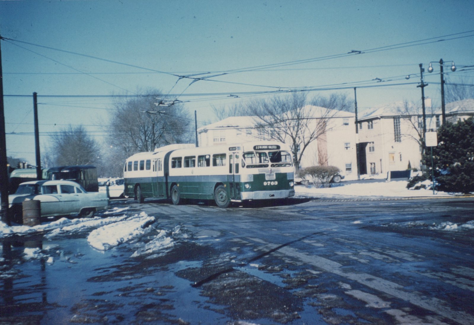 A green and cream trolleybus numbered 9763 is parked on a wet, snowy street under overhead wires, with apartment buildings and bare trees in the background.