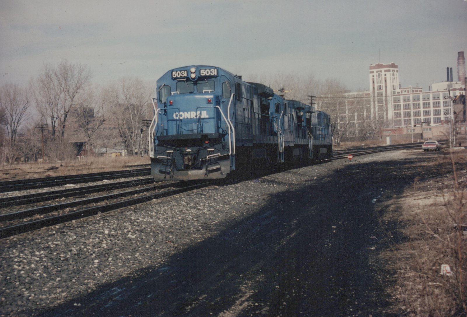 A blue Conrail diesel locomotive numbered 5031 is seen from a three-quarter front view, pulling two other locomotives on a set of tracks, with a white building and bare trees in the background.
