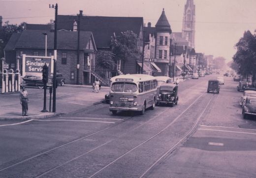 A vintage black-and-white photograph of a Chicago Transit Authority bus from the 1950s on a city street, with embedded trolley tracks, classic cars, and a Sinclair gas station in the background.