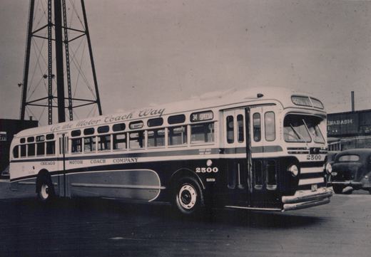 A black and white photo of a Chicago Motor Coach Company bus number 2500, a Flxible New Look model, parked on a street with a tall tower and a freight car in the background.