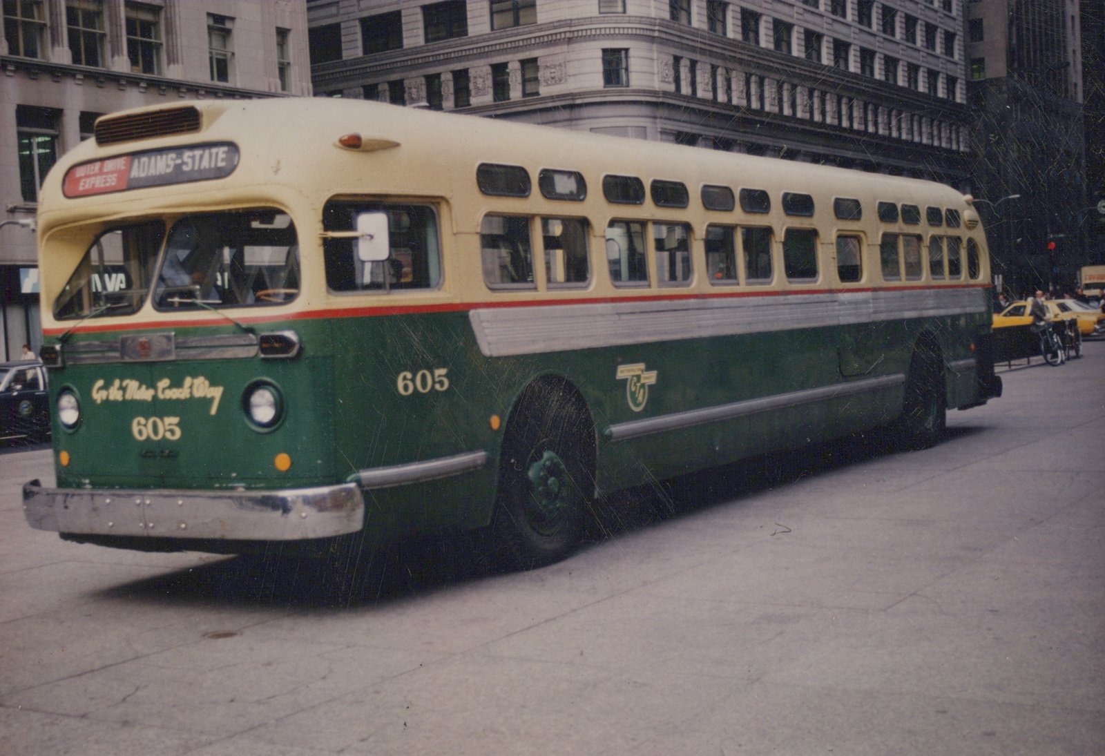 A green and cream Chicago Transit Authority bus, number 605, parked on a city street with tall buildings in the background and a yellow taxi visible.