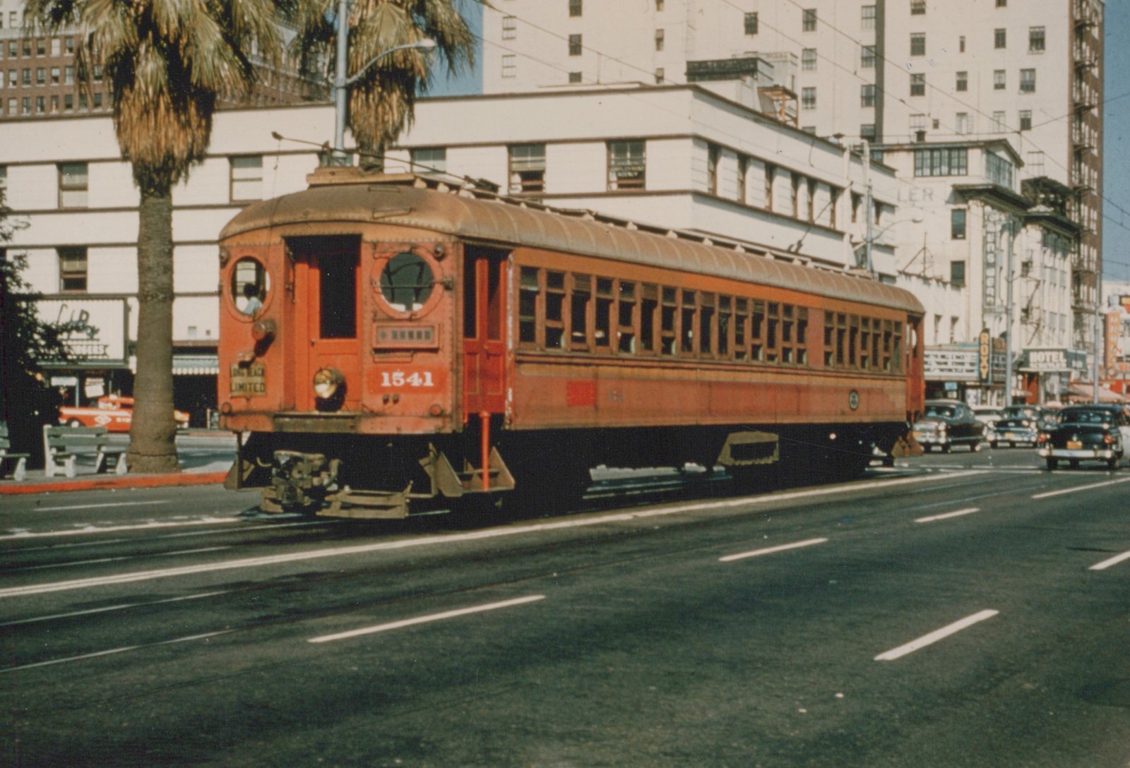 A vintage red electric interurban streetcar on a city street, surrounded by 1950s cars, palm trees, and multi-story buildings.