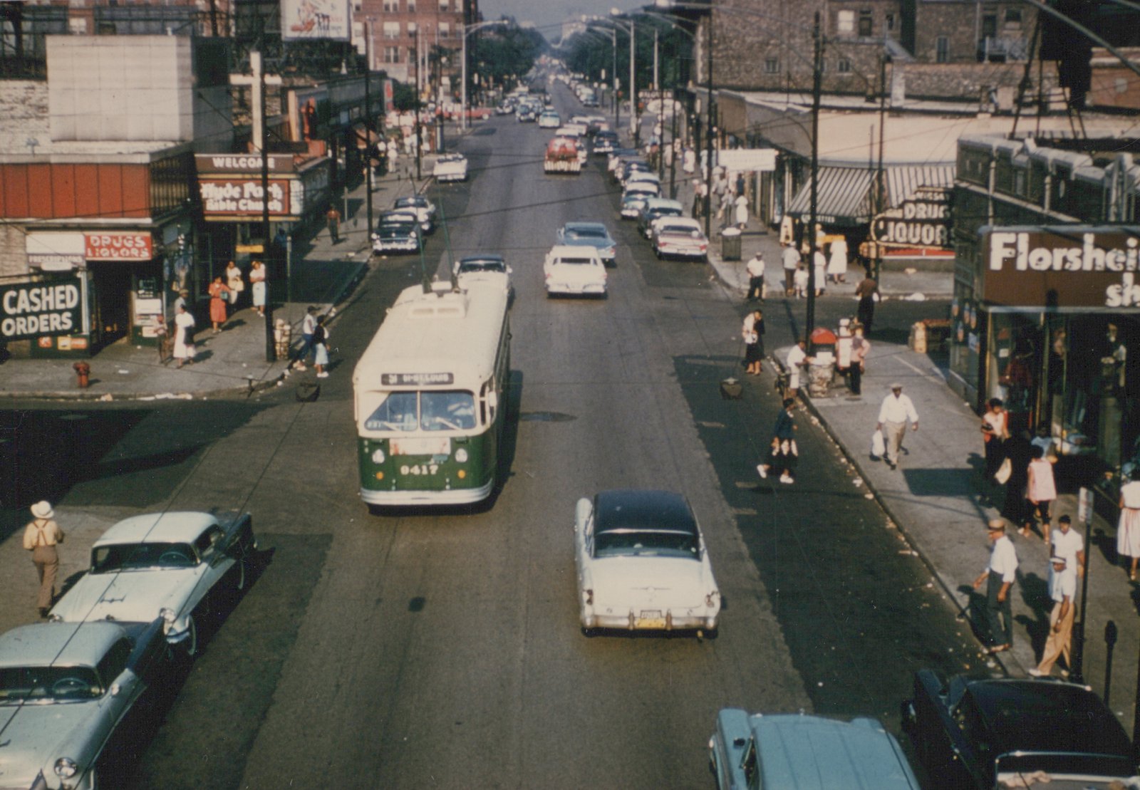 A green and cream trolleybus with the number 9417 is driving down a wide city street lined with 1950s cars and pedestrians on the sidewalks.