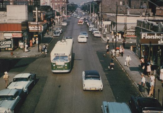A green and cream trolleybus with the number 9417 is driving down a wide city street lined with 1950s cars and pedestrians on the sidewalks.