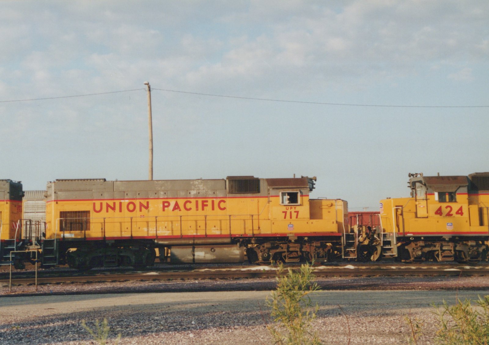 Two yellow Union Pacific diesel locomotives, numbered 717 and 424, are parked side-by-side on tracks in a rail yard with a clear sky and some foliage in the foreground.