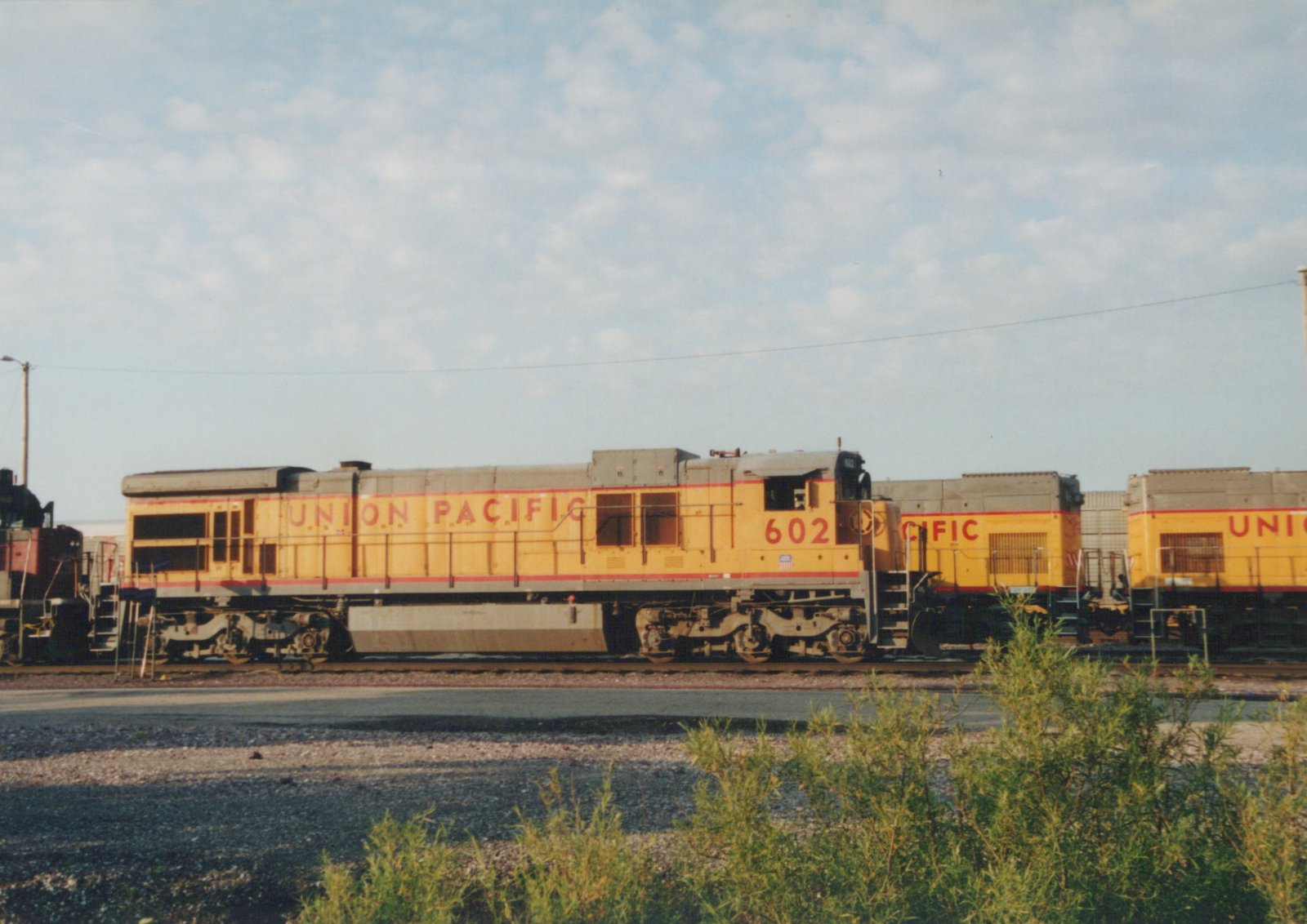A yellow and red Union Pacific diesel locomotive numbered 602 is shown in a side profile at a rail yard, with two more matching locomotives behind it under a partly cloudy sky.