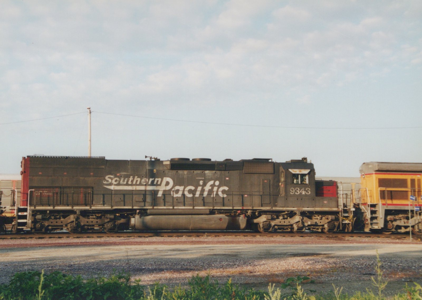 A dark gray Southern Pacific diesel locomotive with the number 9343 on its cab side, parked on tracks in a yard with a yellow locomotive behind it under a partly cloudy sky.