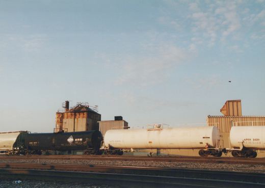 Three tank cars, one black and two white, sit on railroad tracks in a yard with industrial buildings in the background under a clear sky.