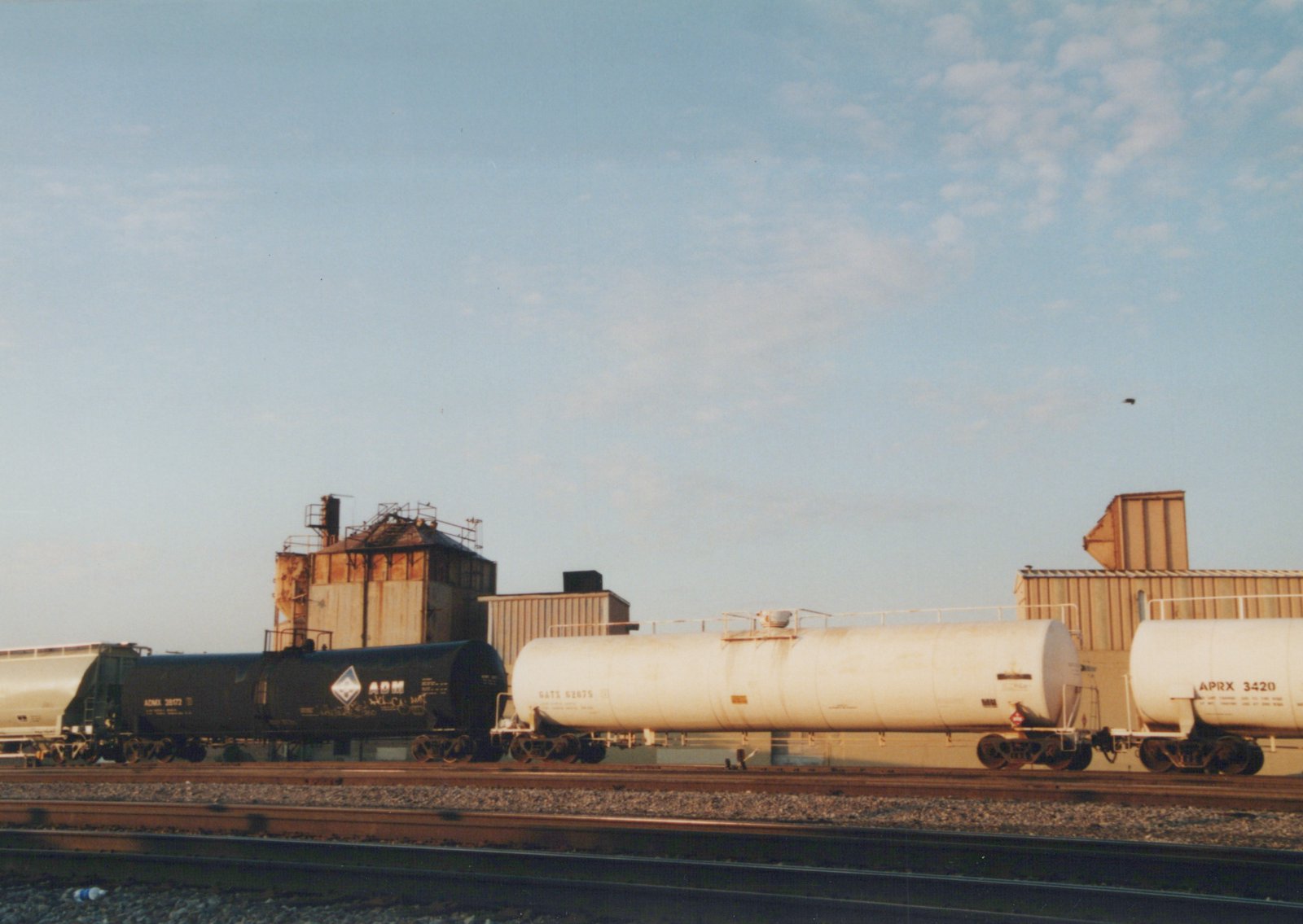 Three tank cars, one black and two white, sit on railroad tracks in a yard with industrial buildings in the background under a clear sky.