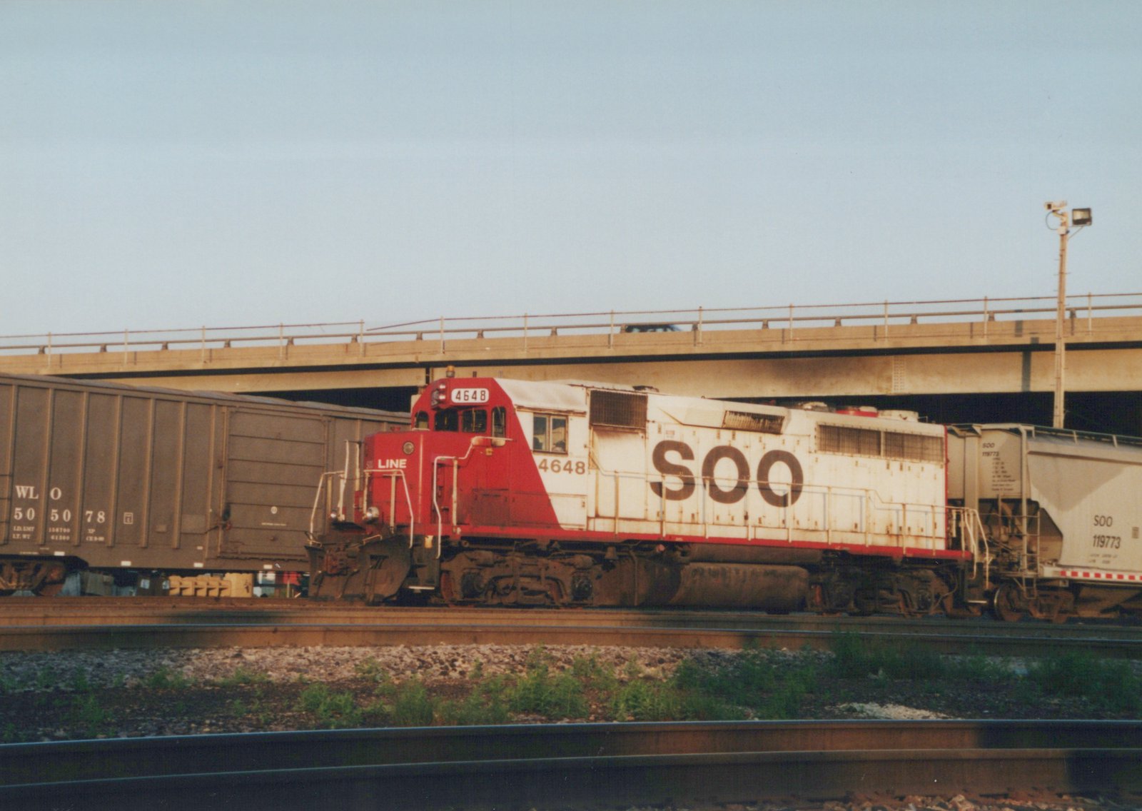 A red and white Soo Line diesel locomotive numbered 4648 pulls freight cars on a railroad track beneath a concrete highway overpass on a sunny day.