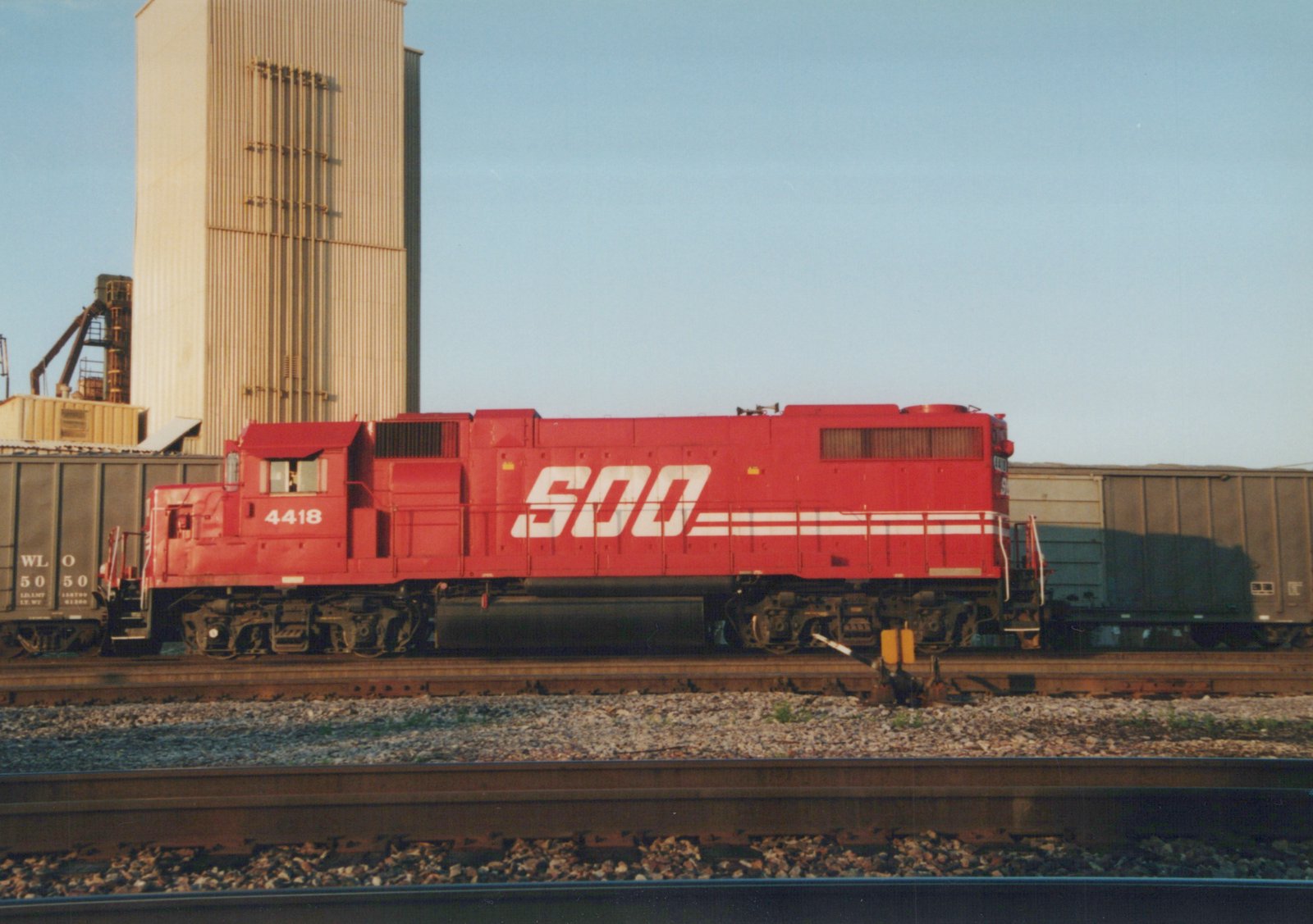 Side view of a red SOO Line diesel locomotive numbered 4418, coupled to freight cars on a set of tracks with a tall silo in the background.