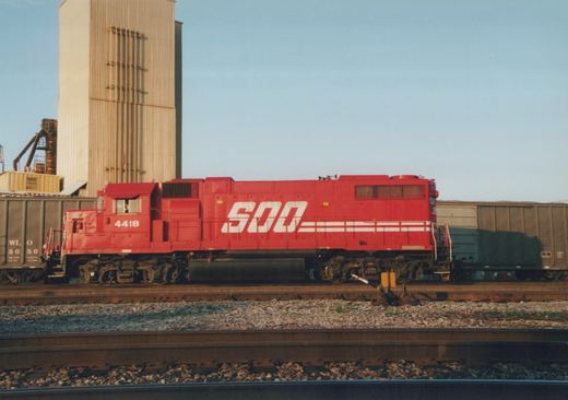 Side view of a red SOO Line diesel locomotive numbered 4418, coupled to freight cars on a set of tracks with a tall silo in the background.