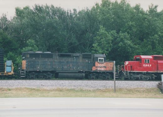 Two diesel locomotives, one black with an orange front and the other solid red, sit on parallel tracks with trees behind them.