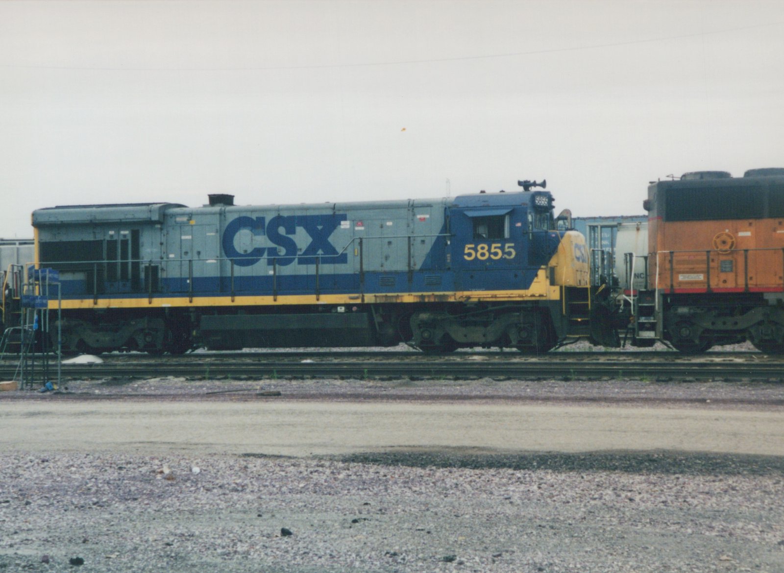 Side view of a blue, gray, and yellow CSX diesel locomotive numbered 5855, parked in a gravel rail yard beside other freight cars.