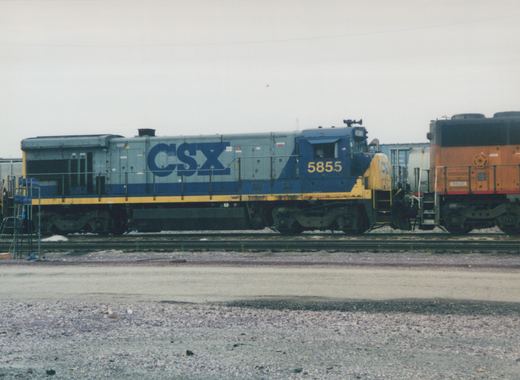 Side view of a blue, gray, and yellow CSX diesel locomotive numbered 5855, parked in a gravel rail yard beside other freight cars.