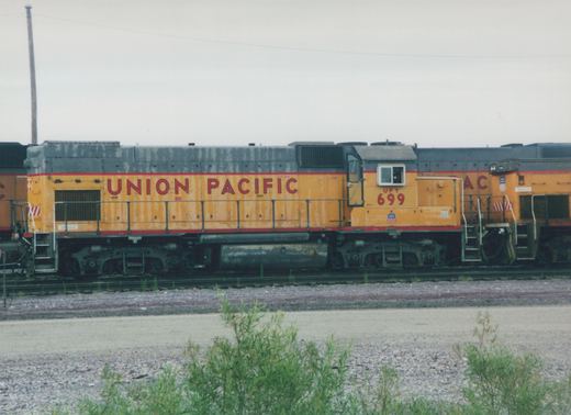 Side view of a yellow and gray Union Pacific diesel locomotive, number 699, sitting on tracks in a gravel yard under an overcast sky.