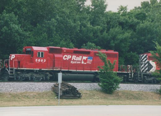 A red CP Rail System diesel locomotive numbered 5683 leads another locomotive on a track beside a road, with trees in the background.