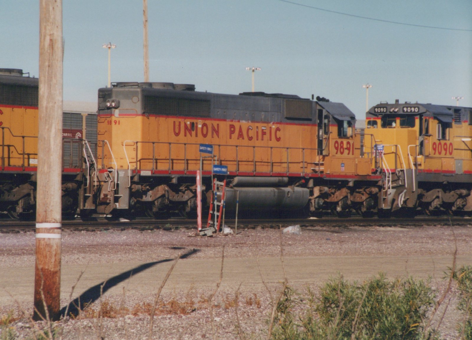 Two yellow and grey Union Pacific diesel locomotives, numbers 9891 and 9090, standing in a rail yard with safety signs nearby and a wooden utility pole in the foreground.