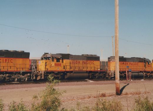 Three yellow and red Union Pacific diesel locomotives are lined up on tracks in a gravel yard on a clear day, with a wooden utility pole in the foreground.