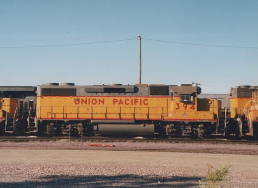 Side view of a yellow and red Union Pacific diesel locomotive, number 394, parked on tracks in a gravel yard with another locomotive nearby.