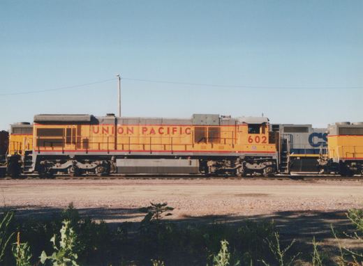 A yellow and grey Union Pacific diesel locomotive, number 602, is shown from the side in a railroad yard under a clear blue sky, with a grey CSX locomotive visible on an adjacent track.