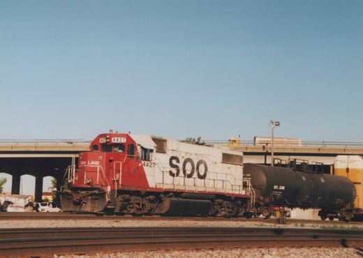 A red and white SOO Line diesel locomotive numbered 4427 is pulling freight cars on railroad tracks under a concrete bridge on a sunny day, with a white van parked nearby and a yellow truck visible on the bridge above.