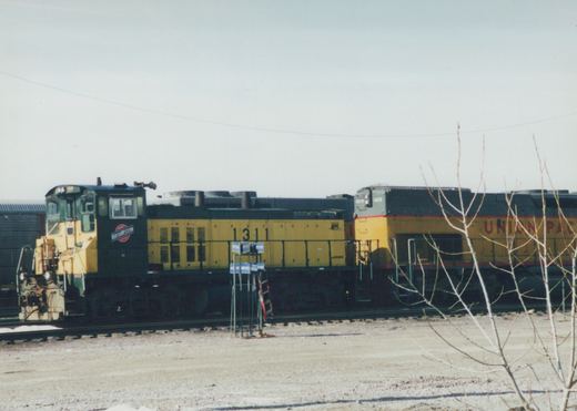 Side view of two yellow and black diesel locomotives, one marked Chicago and North Western 1311, coupled in a gravel yard with bare trees and snow patches under an overcast sky.